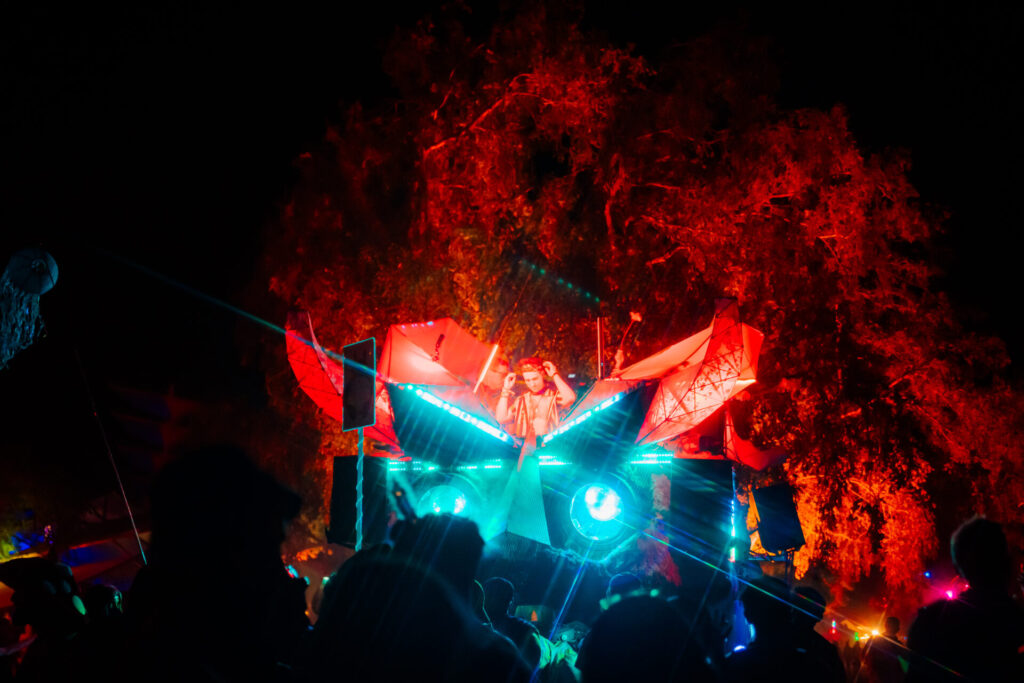 A fully illuminated monster-themed art car with glowing eyes hosts a late night DJ set.